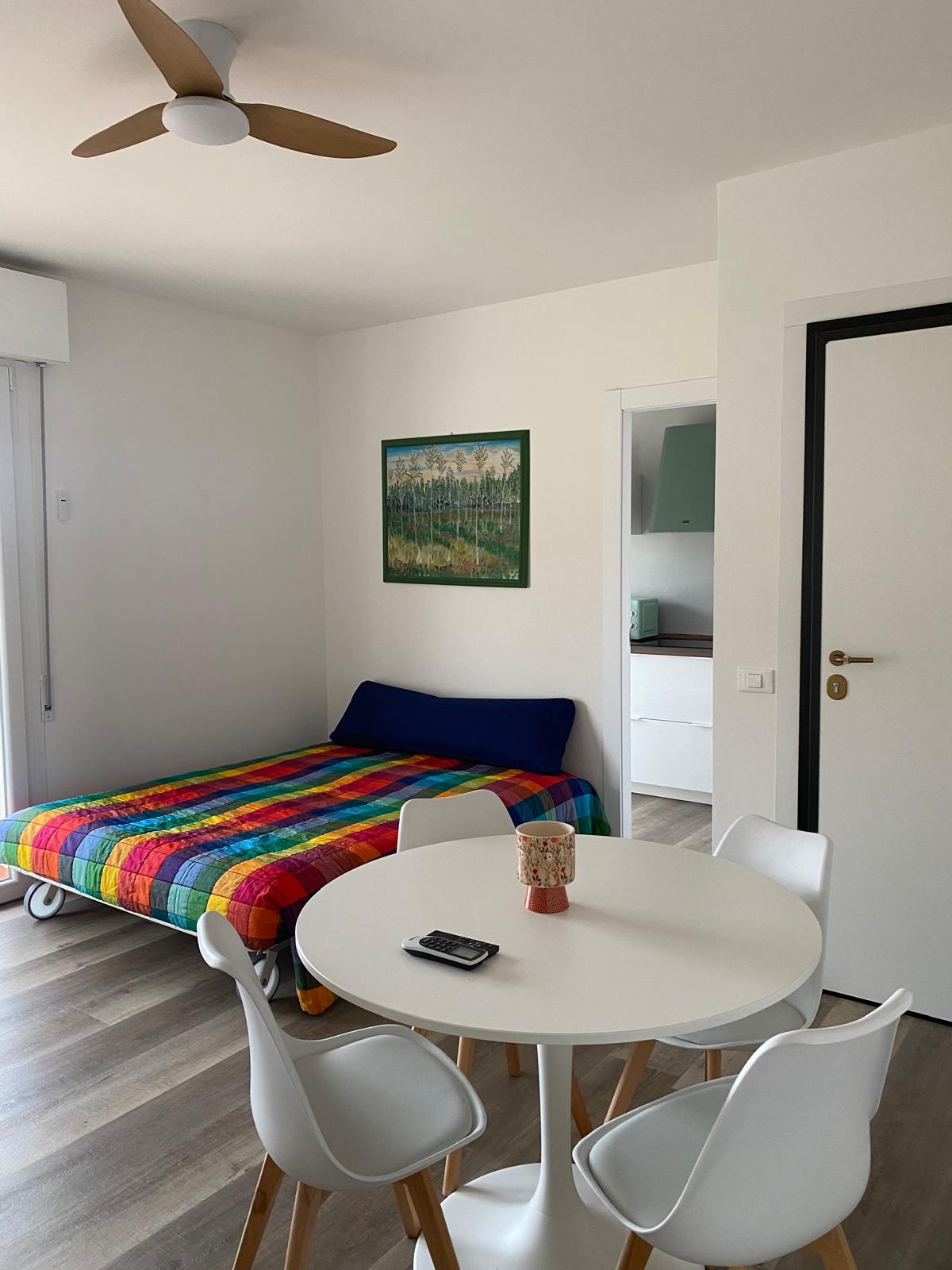 Soggiorno della Maison de Gio' con divano letto a quadri arcobaleno, tavolo rotondo bianco e cucina verde acqua sullo sfondo