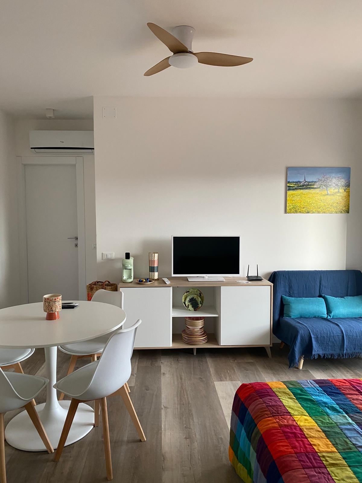 Soggiorno di La Maison de Gio' con tavolo bianco rotondo, divano blu, TV, climatizzatore, ventilatore e copriletto arcobaleno
