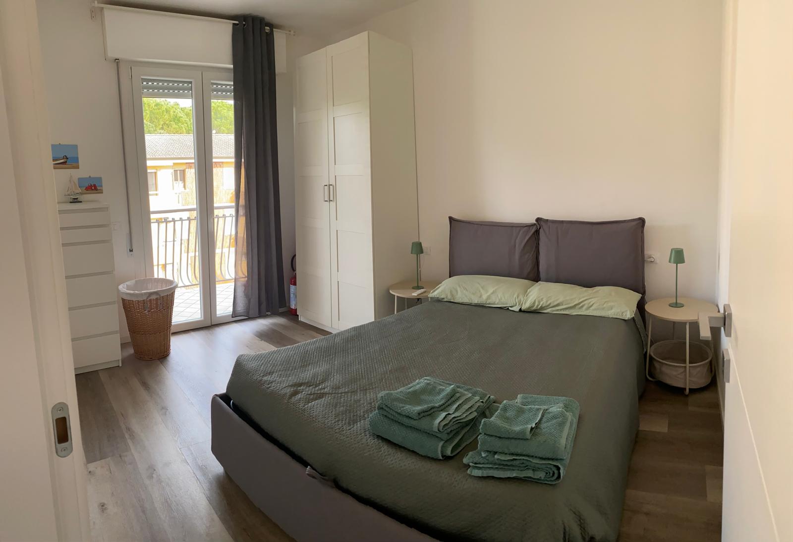 Camera de La Maison de Gio' con letto matrimoniale grigio, copriletto verde, asciugamani e portafinestra sul balcone