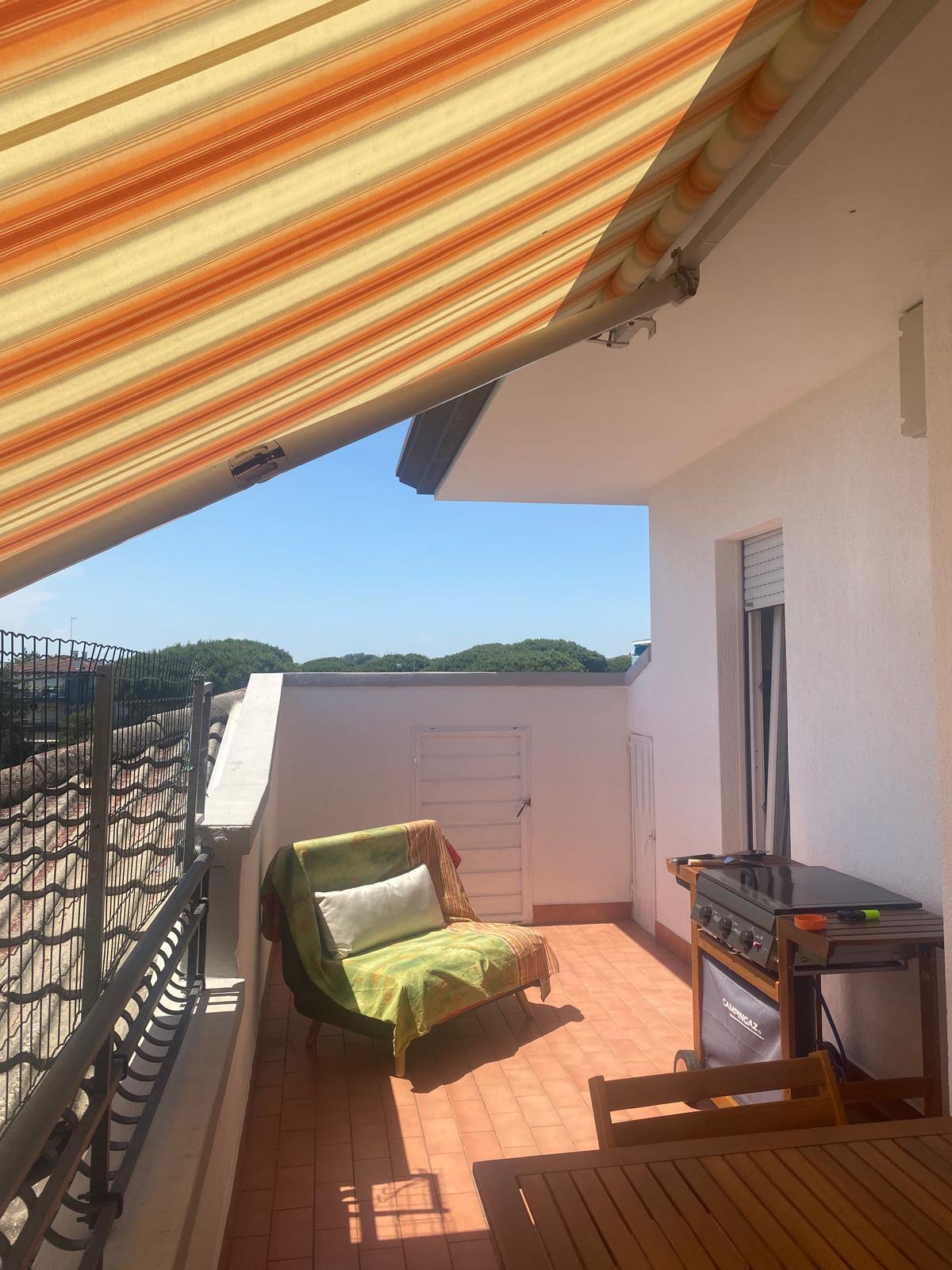 Terrazza in cotto con tenda a righe arancioni, poltrona, barbecue Campingaz, tavolo in legno e pini sullo sfondo